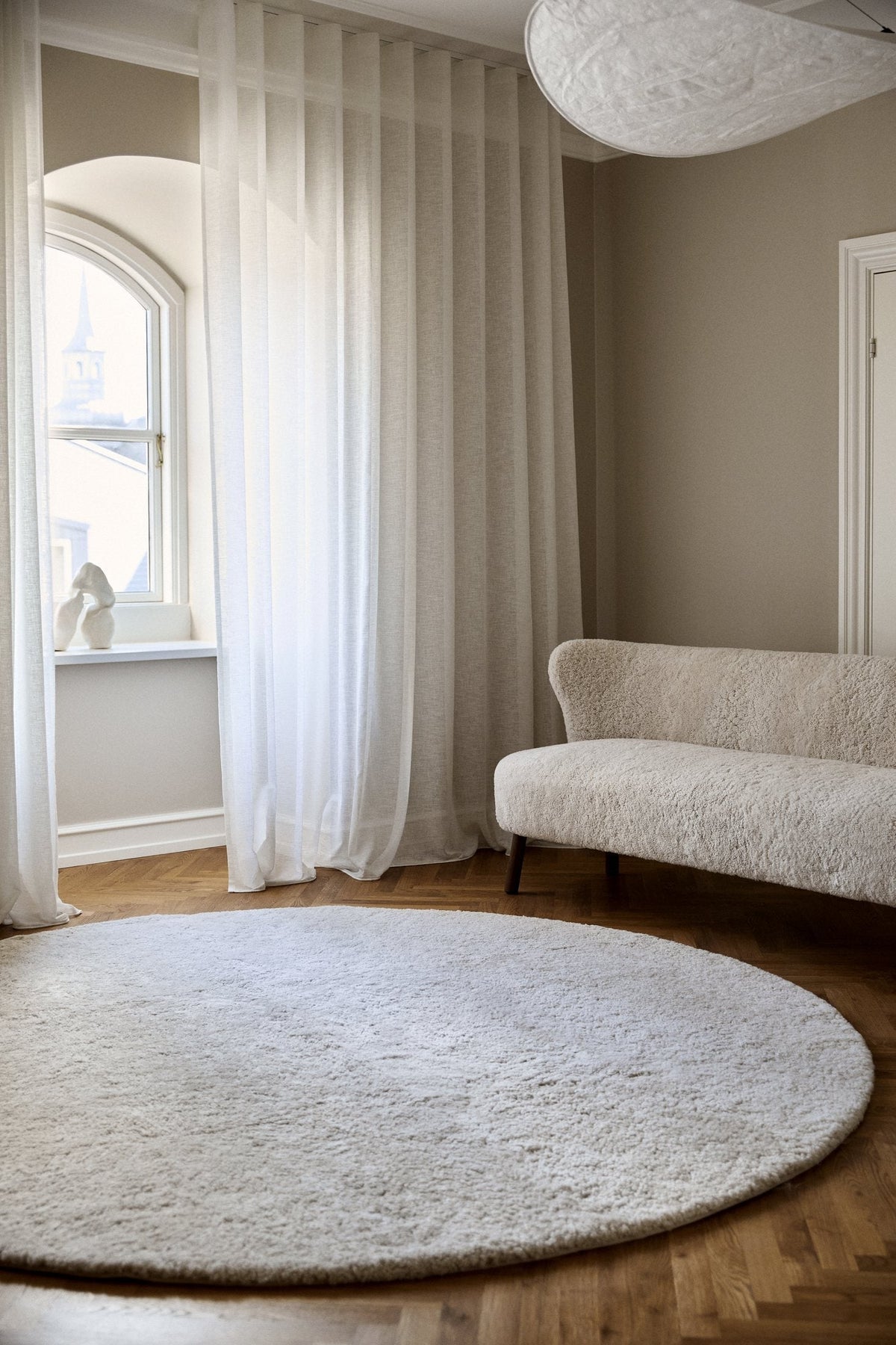Round white short-wool sheepskin rug in a bright room with sheer curtains and a curved sofa